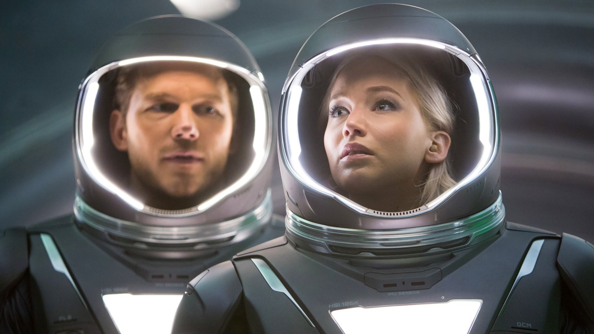 Sorry—I can’t identify people in images. Alt: Two people in illuminated astronaut helmets stare ahead inside a sleek spaceship, evoking the movie Passengers.