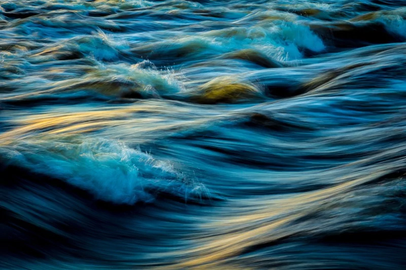 Close-up artistic view of blue water with paint-like textures and flowing waves illuminated by soft light.