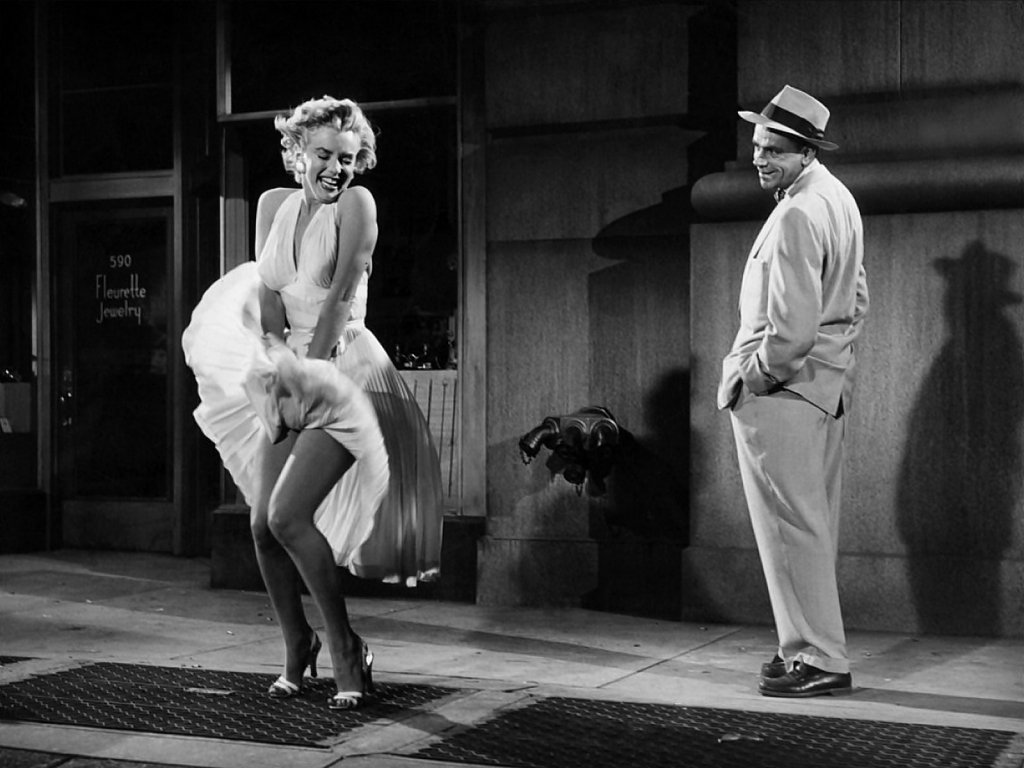 Marilyn Monroe stands on a city street, her white dress billowing around her as a man in a light suit watches, creating a classic Hollywood scene filled with glamour and charm.