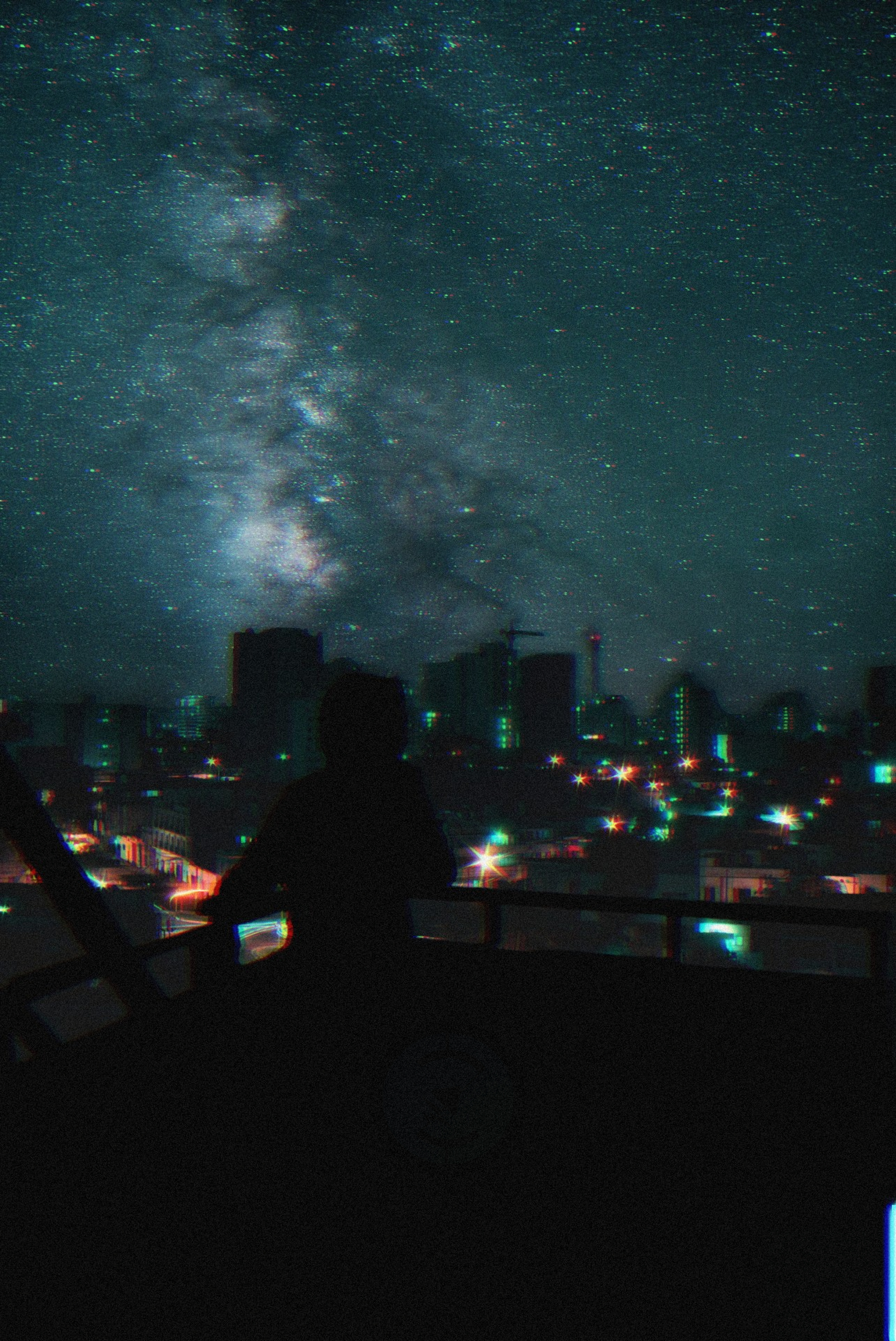 Silhouette on a balcony gazing at a bright, star-filled night sky and Milky Way above a man-made cityscape.