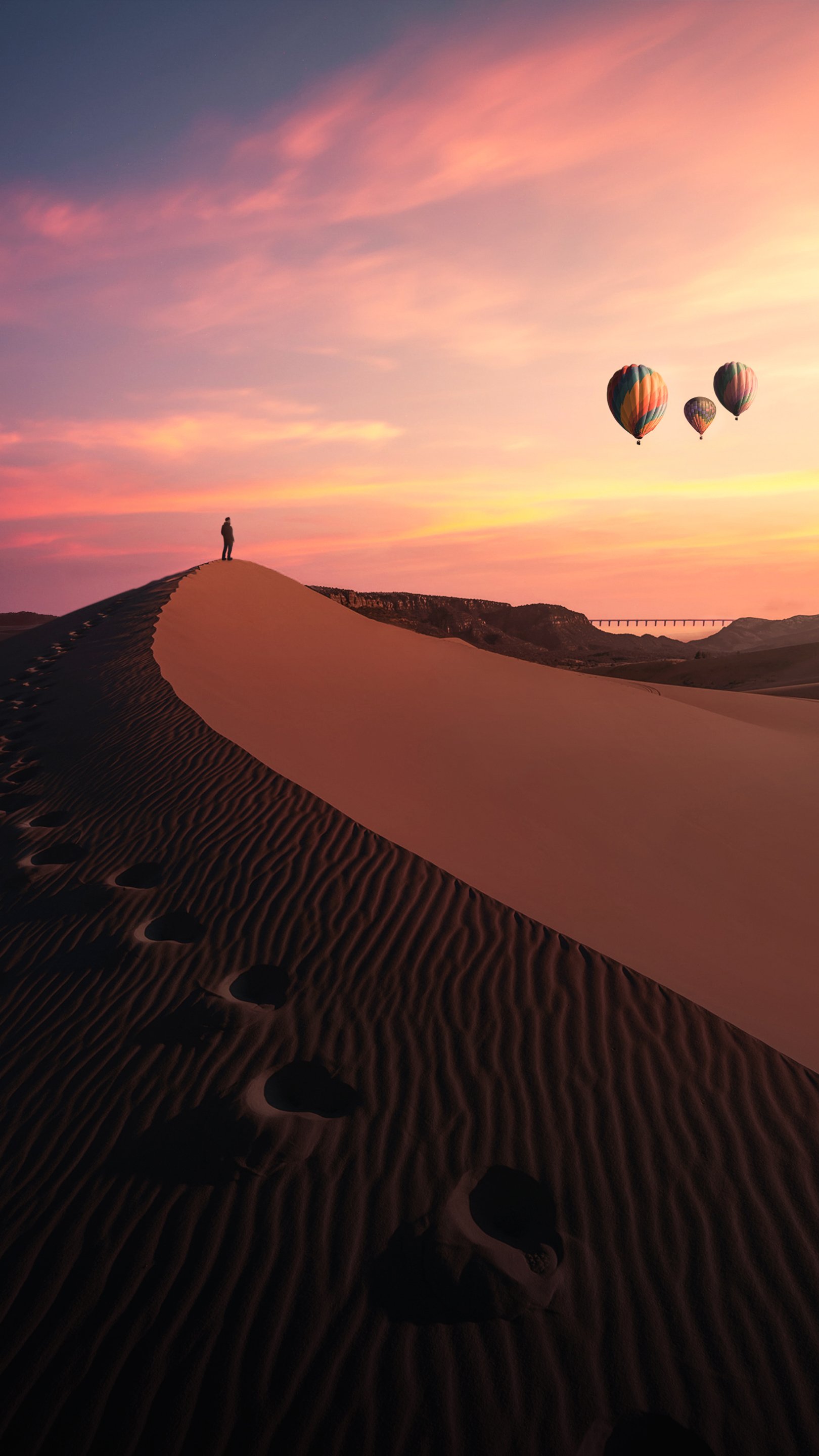 Artistic desert scene: lone figure atop a sand dune, rippling sand and footprints below, colorful hot-air balloons drifting across a pastel sunset.