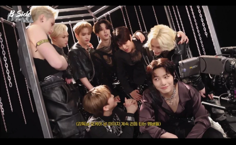 K-pop performers in black leather and chains pose closely on a Venom-themed set, playful expressions amid hanging chains, studio lights and a camera visible.