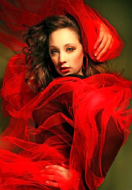 A woman poses dramatically in flowing red fabric, showcasing an artistic expression that emphasizes movement and emotion against a soft green backdrop.