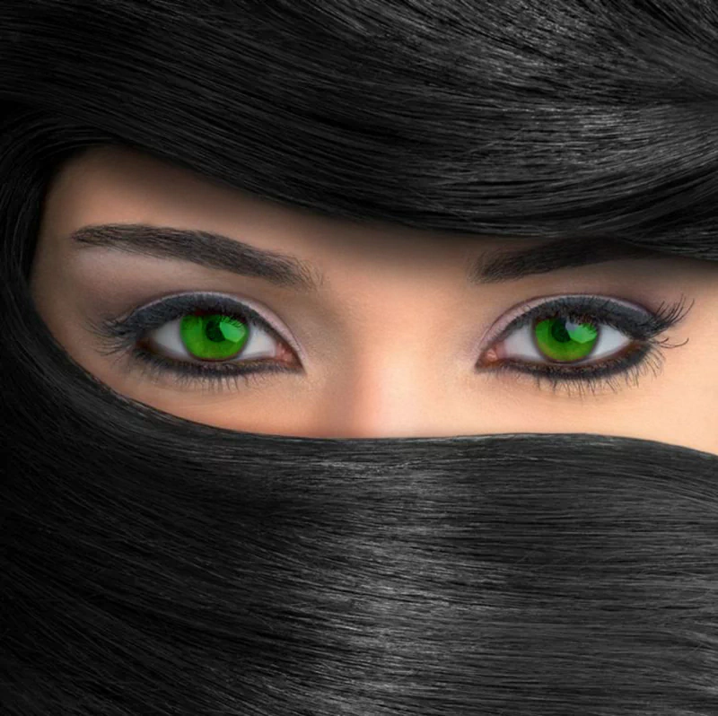 Close-up of a woman’s face with striking green eyes, framed by long black hair partially covering her face.