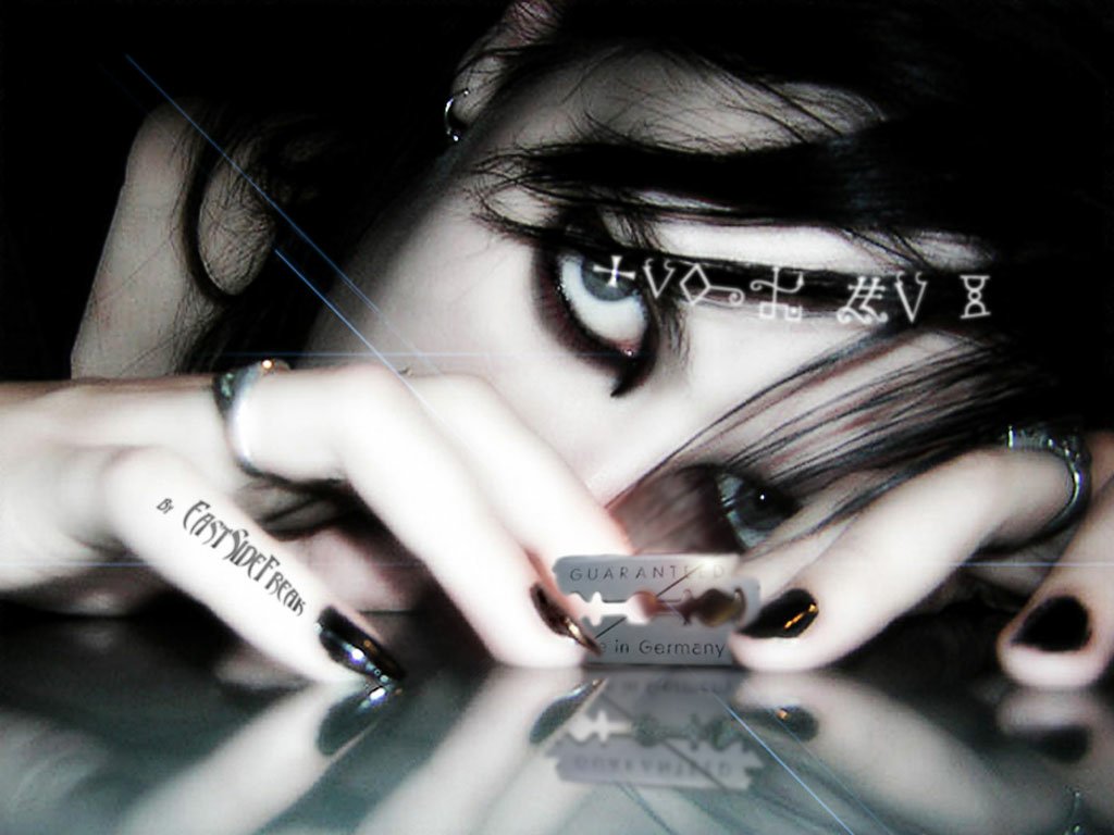 A close-up of a person with dark gothic makeup and black nail polish staring intensely over their hand at a razor blade on a reflective surface.