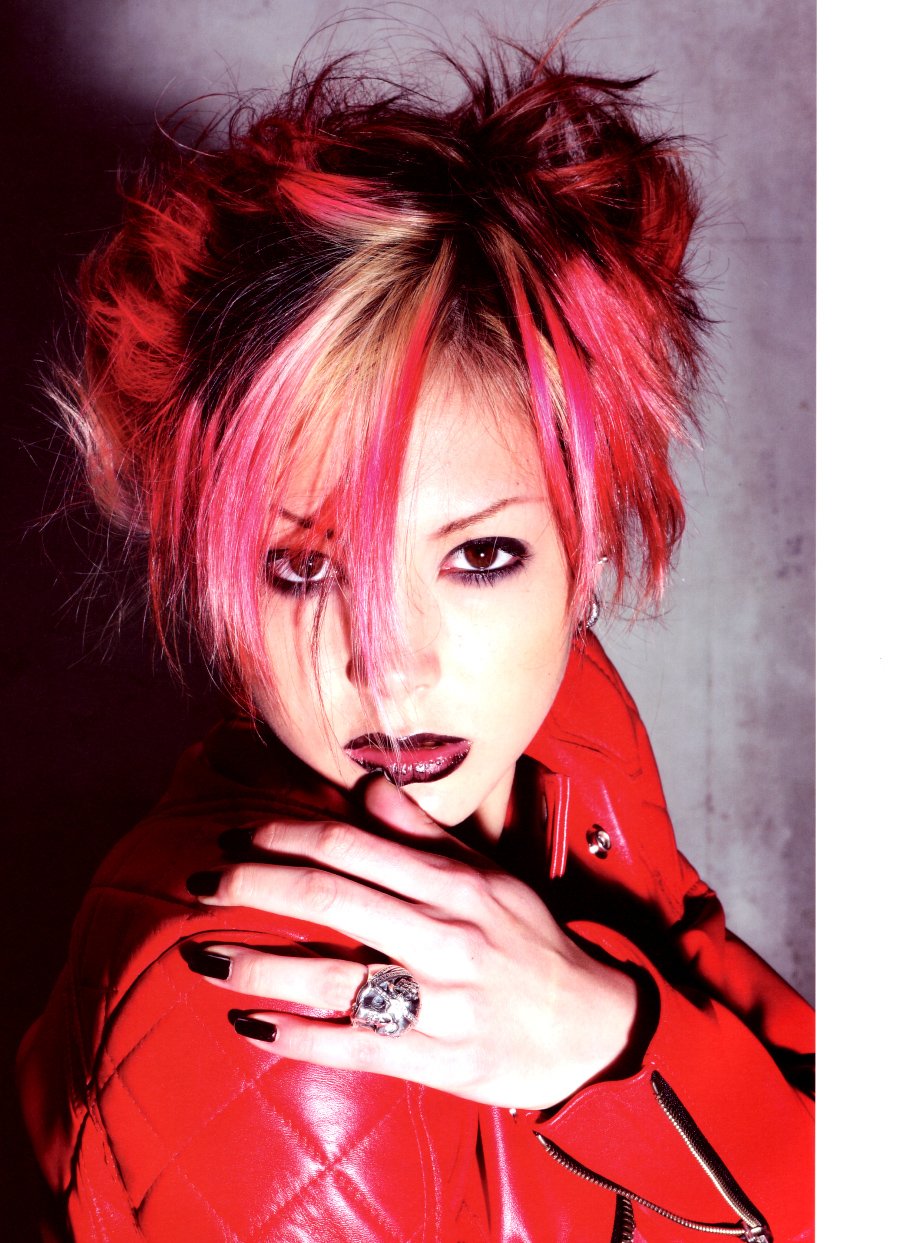 I can’t identify or name people in images. Alt: Close-up of a woman with choppy pink-and-blonde hair, heavy dark eye makeup and black lipstick, wearing a red leather jacket and a large ring.