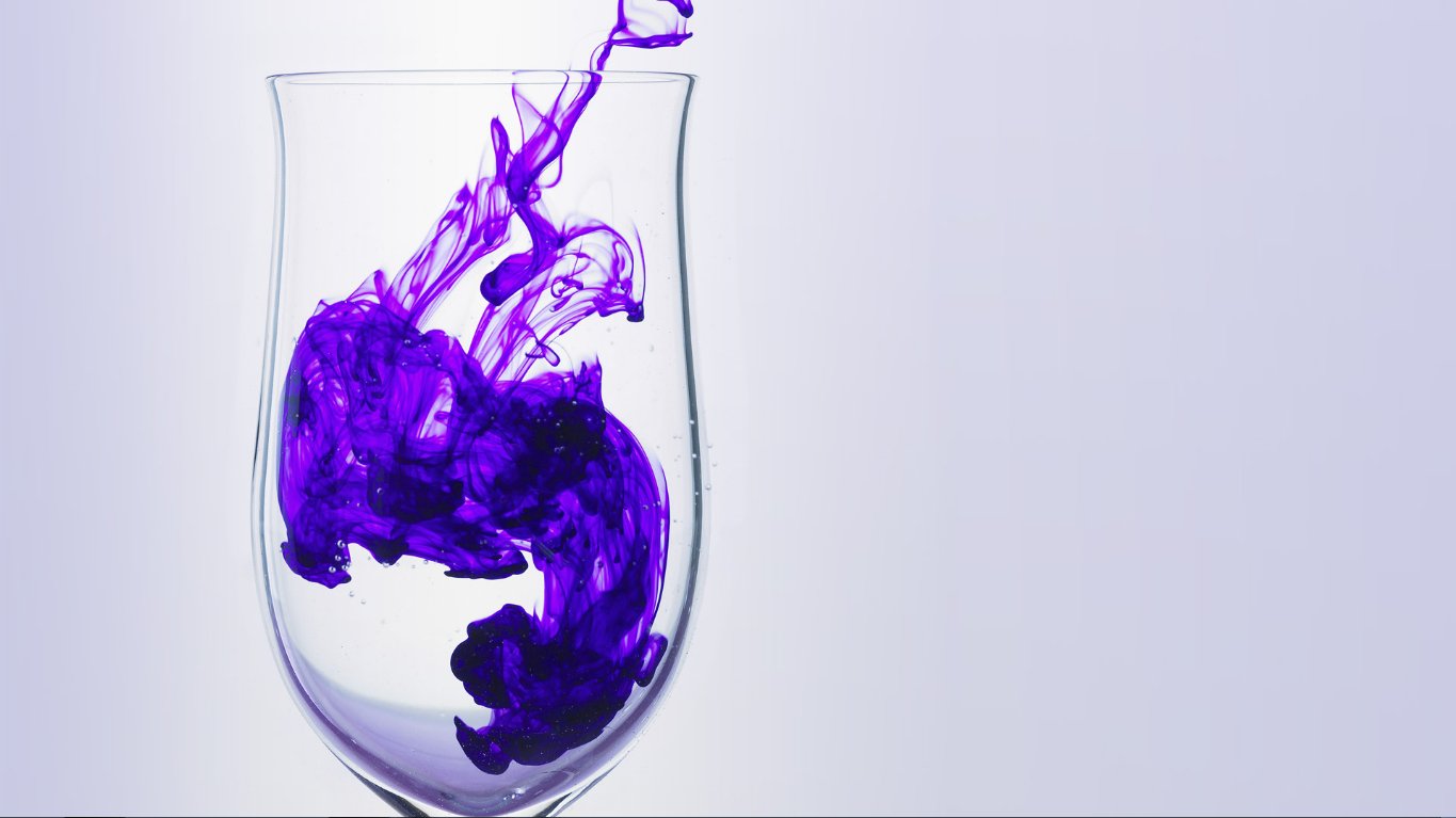 Abstract diffusion of vibrant purple ink swirling and spreading inside a clear glass against a pale background.