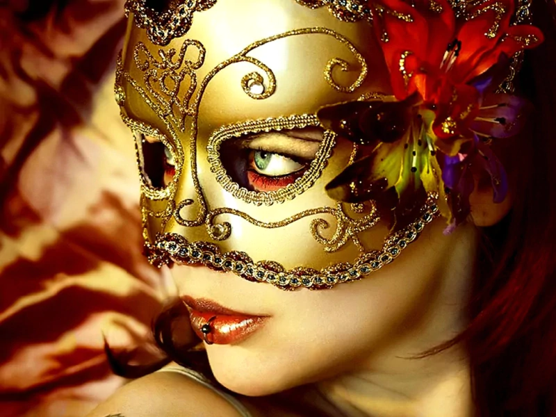 Artistic portrait of a woman wearing an ornate gold carnival mask with red hair and a flower accent, gazing sideways under warm, dramatic lighting.