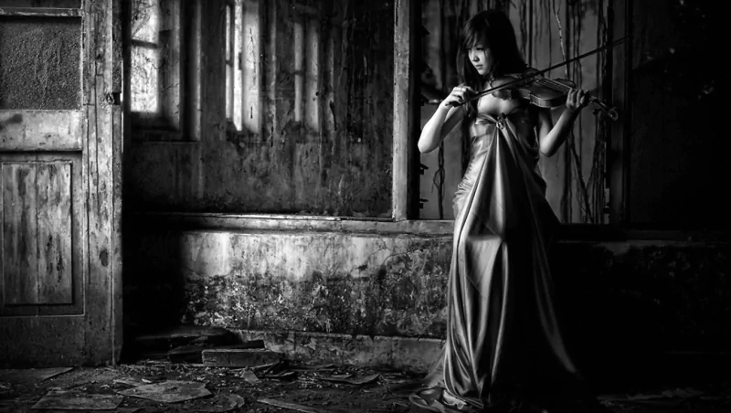 A fantasy-themed black and white image of a woman in a flowing gown playing a violin in a dim, rustic room with textured walls and old doors.