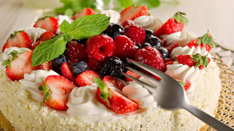 A fresh fruit cake topped with strawberries, raspberries, blackberries, blueberries, and garnished with mint leaves on a woven placemat.