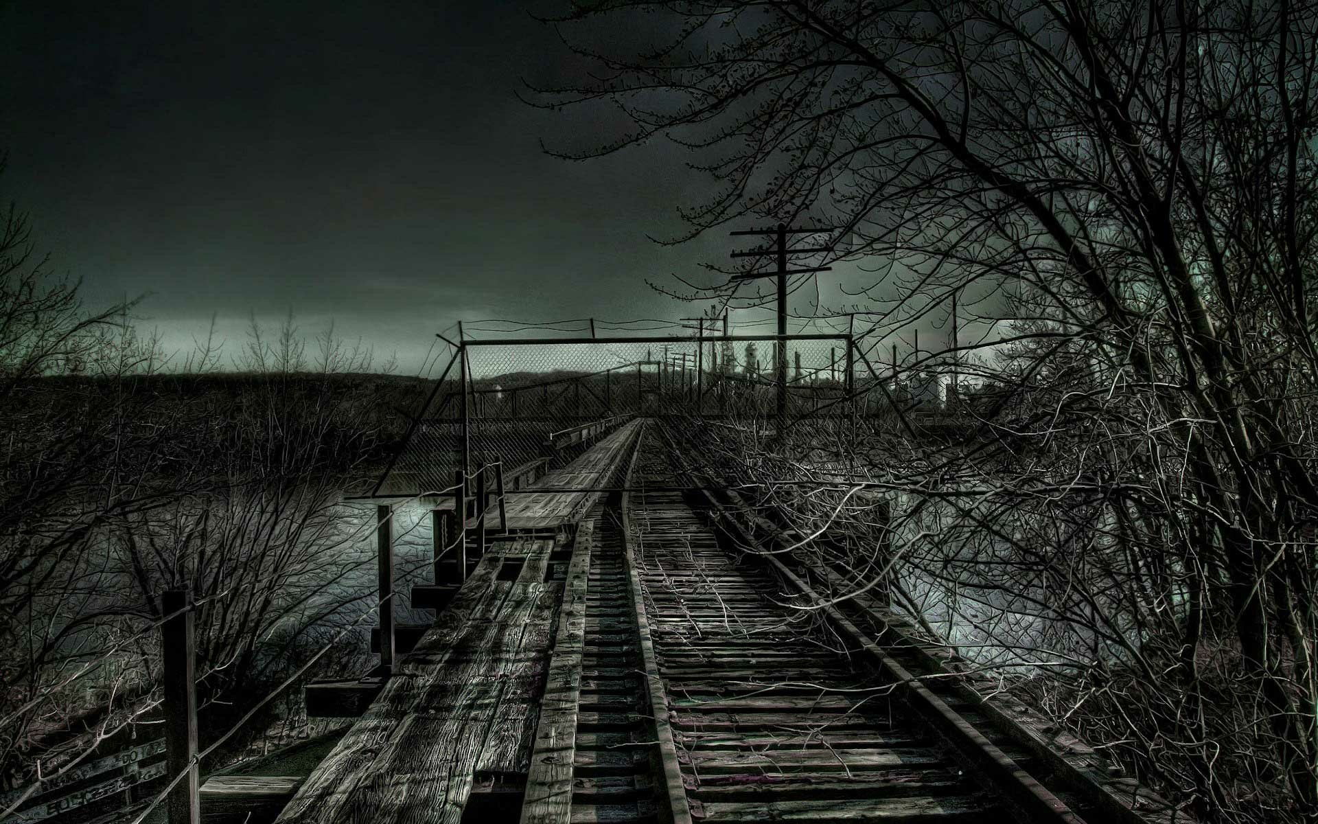 Weathered man-made railroad bridge of wooden planks stretching over water at dusk, framed by leafless trees under a dark, moody sky.