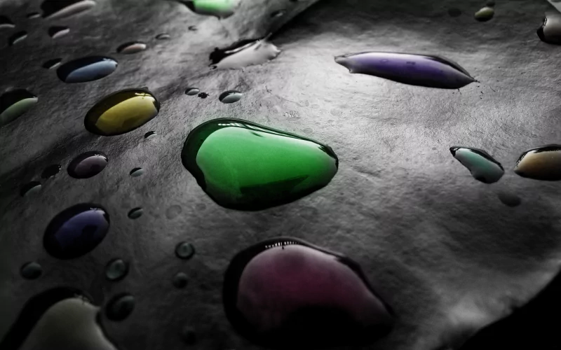 A close-up photograph showcases various water drops in vibrant colors resting on a dark surface, creating a striking contrast and highlighting the beauty of nature’s reflections.