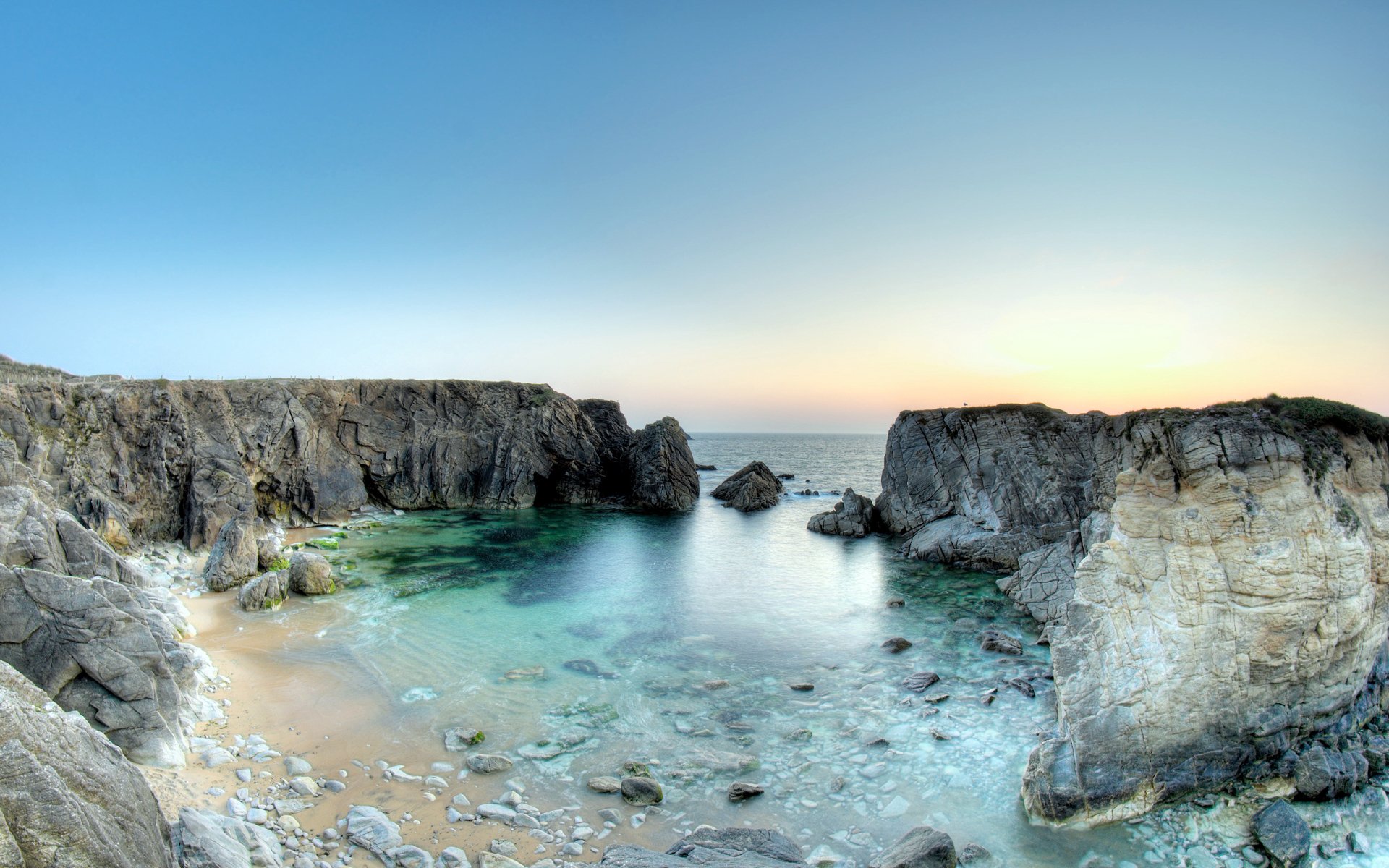 Clear, calm water surrounds rugged rock formations in a tranquil nature setting at sunset.