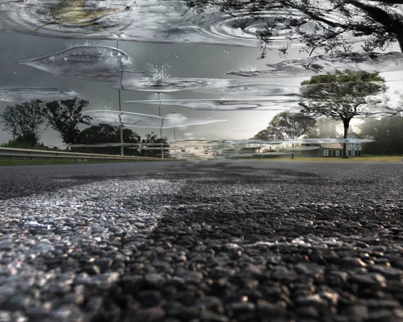 Artistic, rain-soaked low-angle view of wet asphalt with puddle reflections, moody clouds and silhouetted trees and houses lining the distant road.