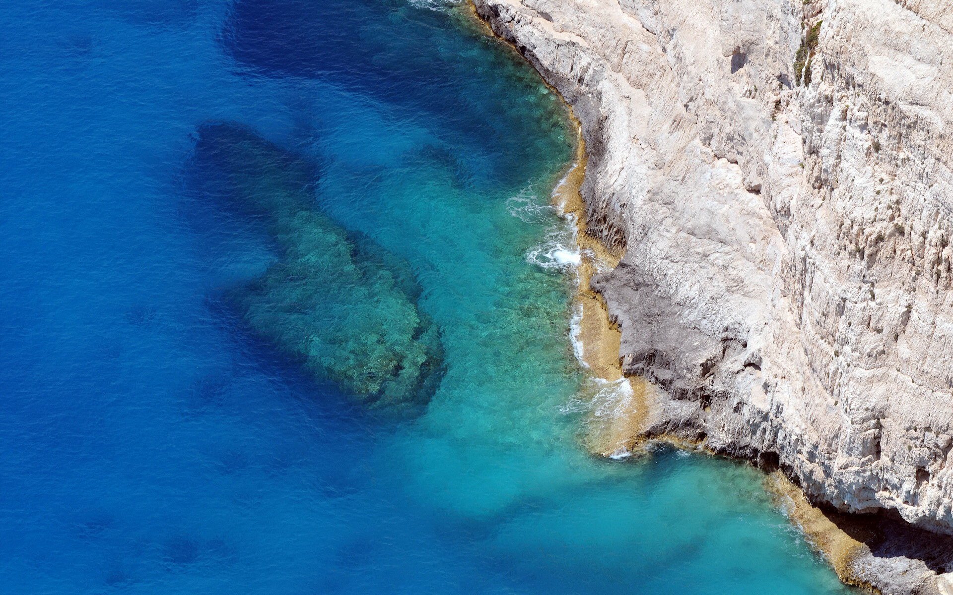 Aerial nature seascape of a rocky coastline with white cliffs and clear turquoise-to-deep-blue water revealing submerged rocks.