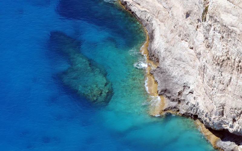 Aerial nature seascape of a rocky coastline with white cliffs and clear turquoise-to-deep-blue water revealing submerged rocks.