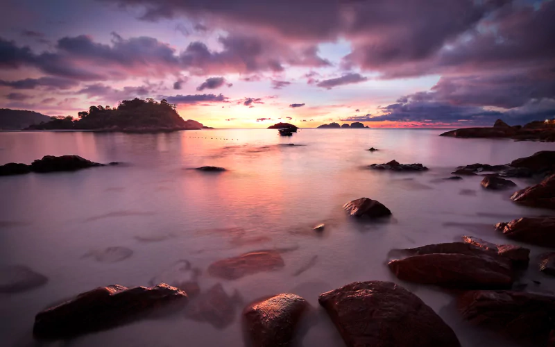 Nature seascape at sunset — smooth water laps dark rocks and distant islets beneath a vivid pink-and-purple sky.