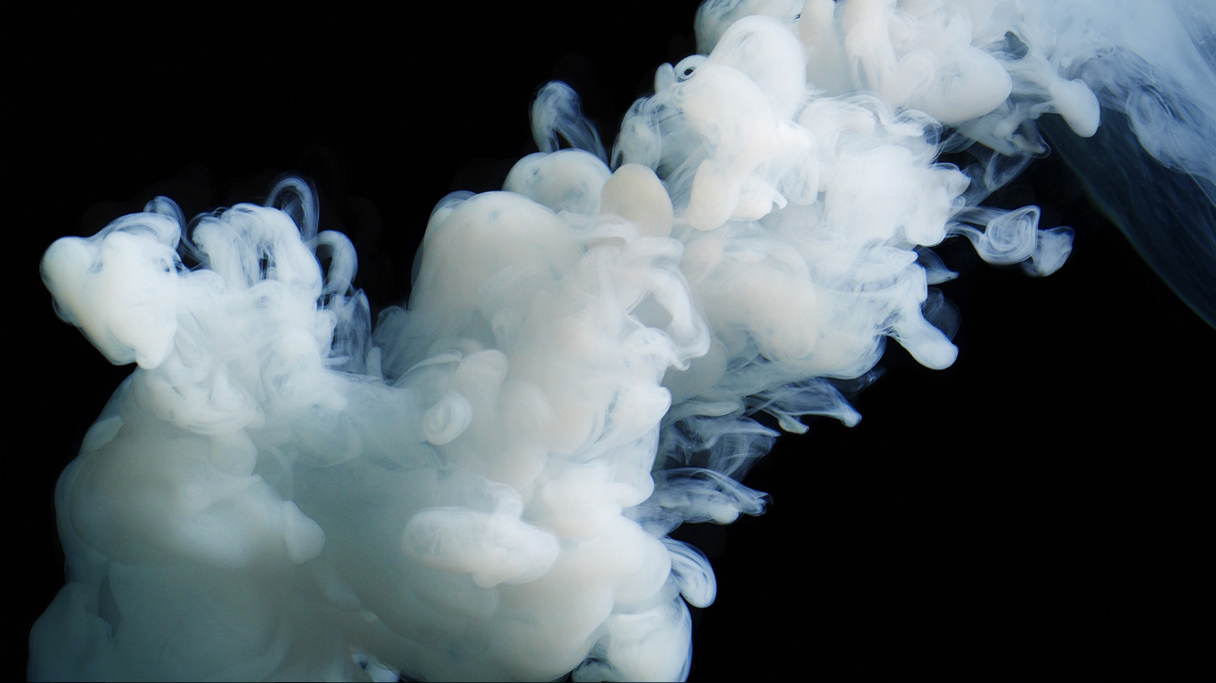 Abstract diffusion of milky white smoke curling and billowing into soft plumes and tendrils against a deep black background.