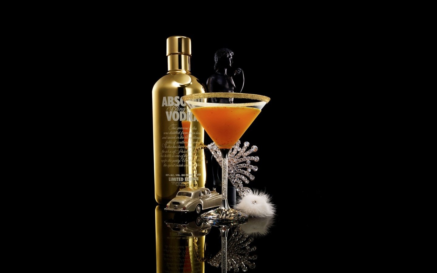 A stylish orange cocktail in a martini glass with decorative accents, set next to a gold Absolut Vodka bottle and a small silver car on a reflective black surface.