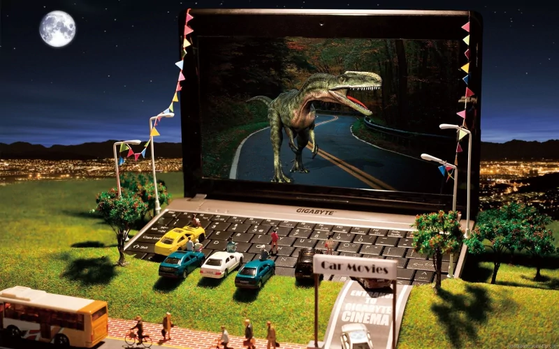 A creatively manipulated photograph features a miniature drive-in scene with a laptop displaying a dinosaur on a road, set against a night sky and city lights.