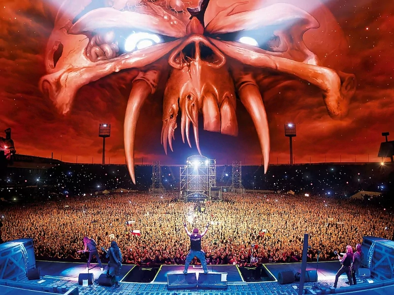 A massive crowd at an Iron Maiden concert with the band's iconic fiery skull artwork dominating the sky above the stage.