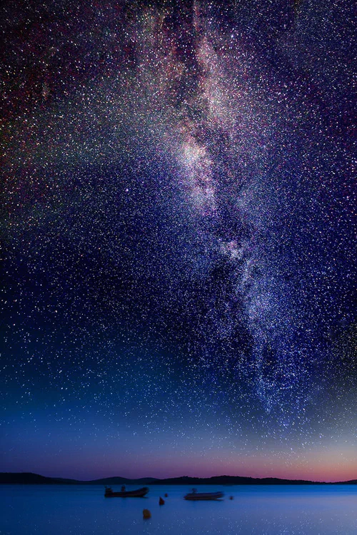 A breathtaking fantasy landscape at night, featuring a star-filled sky with the Milky Way stretching above calm waters and silhouetted boats.