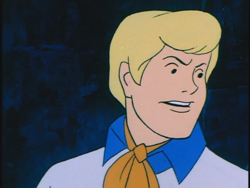 Fred Jones from the TV show Scooby-Doo: blond man in a white shirt and orange ascot looking off-screen against a dark background.