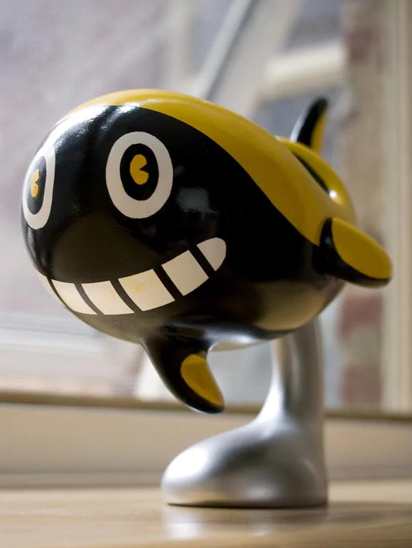 Man-made decorative glossy black-and-yellow sharky whale sculpture with a cartoon grin and big round eyes, mounted on a silver stand.