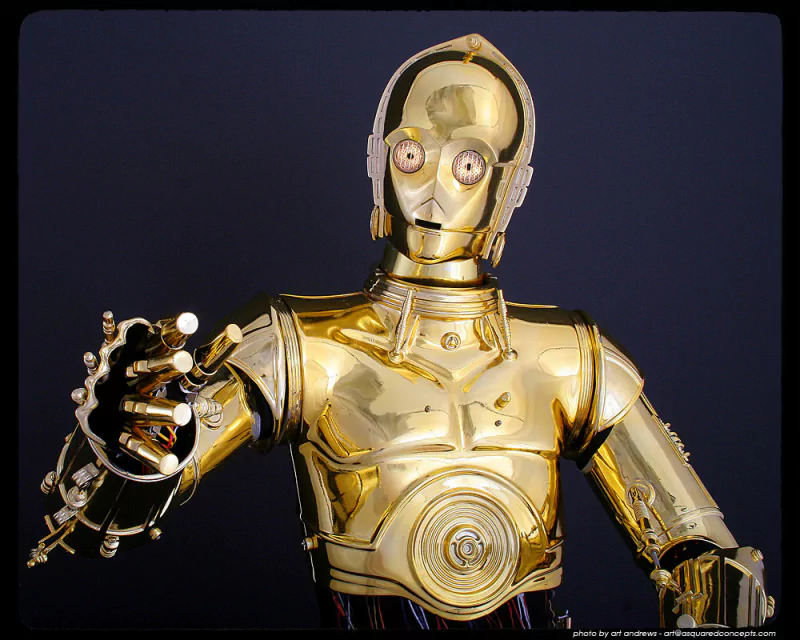 Golden C-3PO robot from the Star Wars movie, shown against a dark background with its arm extended forward.