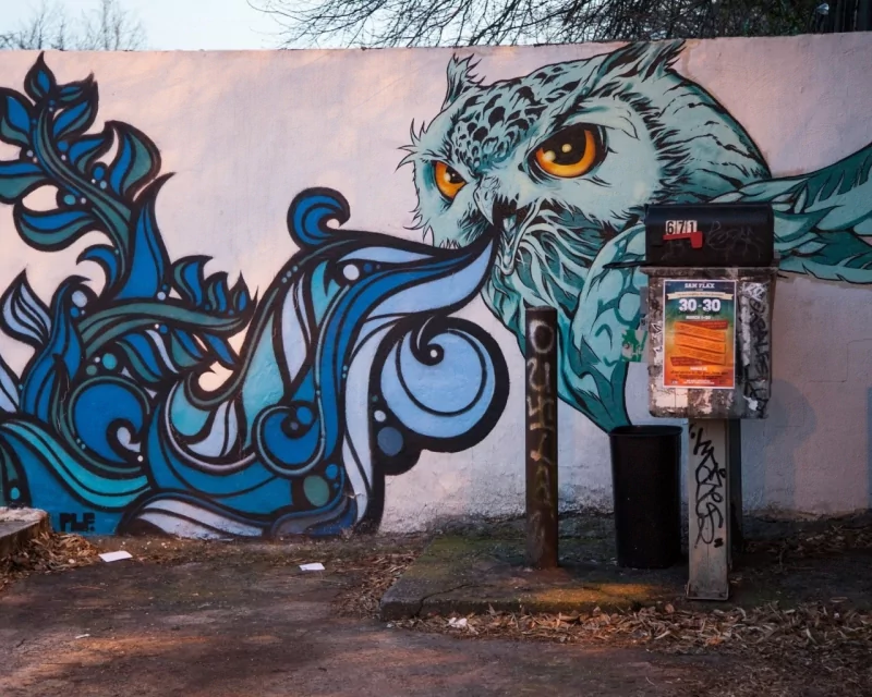Artistic graffiti of a fierce owl with striking orange eyes blowing intricate blue swirls on a white wall beside old, weathered public phones.