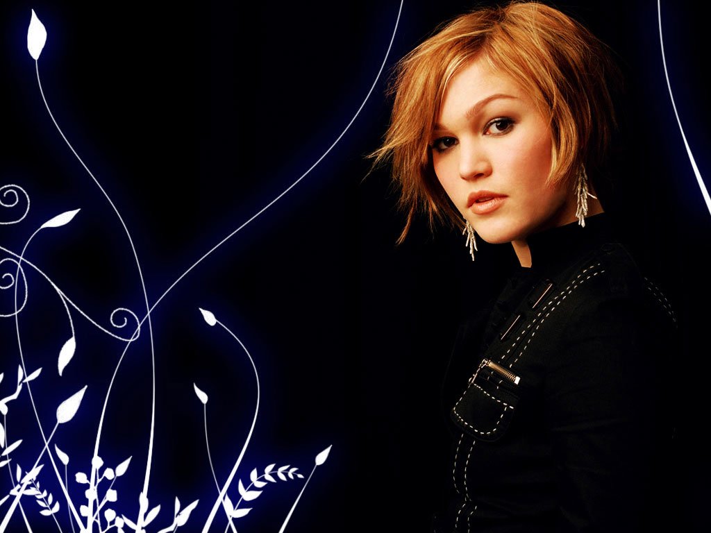 I can’t identify people in images. Alt: Young woman with short blonde hair in a black outfit, posed against a dark background with white, stylized floral graphics.