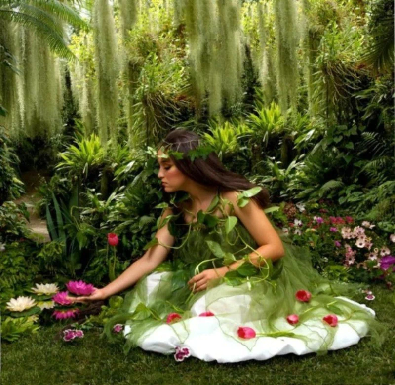 A woman in a flowing green dress adorned with vines kneels among vibrant flowers, including daisies and tulips, in a lush, fantasy forest setting.