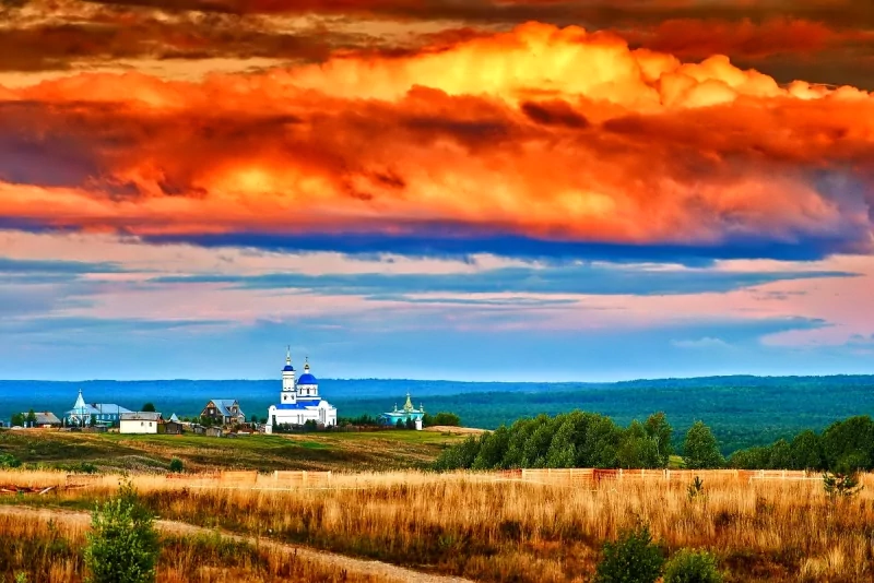 A vibrant nature landscape featuring a golden field, distant white buildings, and a dramatic orange and blue sky at sunset.