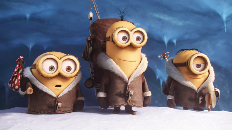 Movie still: three Minions in heavy winter coats stand on snow in an icy cave; one carries a red polka-dot bundle, another holds a small flag.