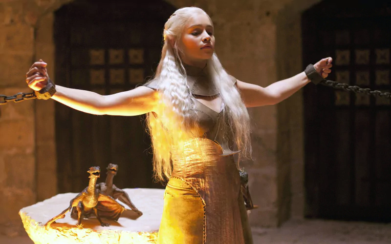 Daenerys Targaryen stands with arms outstretched, chained in a dimly lit stone chamber from the TV show Game of Thrones.