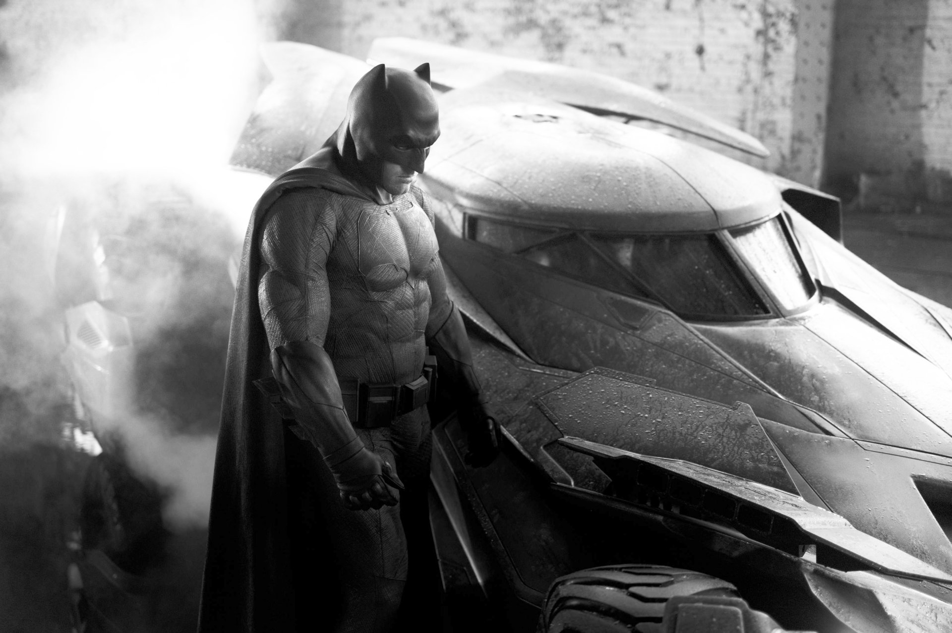 Batman stands next to the Batmobile in a dark, smoky setting from the movie Batman V Superman: Dawn of Justice.