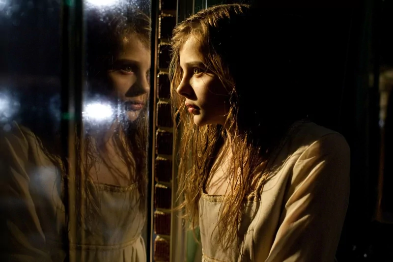 Moody cinematic still evoking Let Me In: a young woman with wet hair peers at her reflection in a rain-streaked window, bathed in warm, shadowed light.