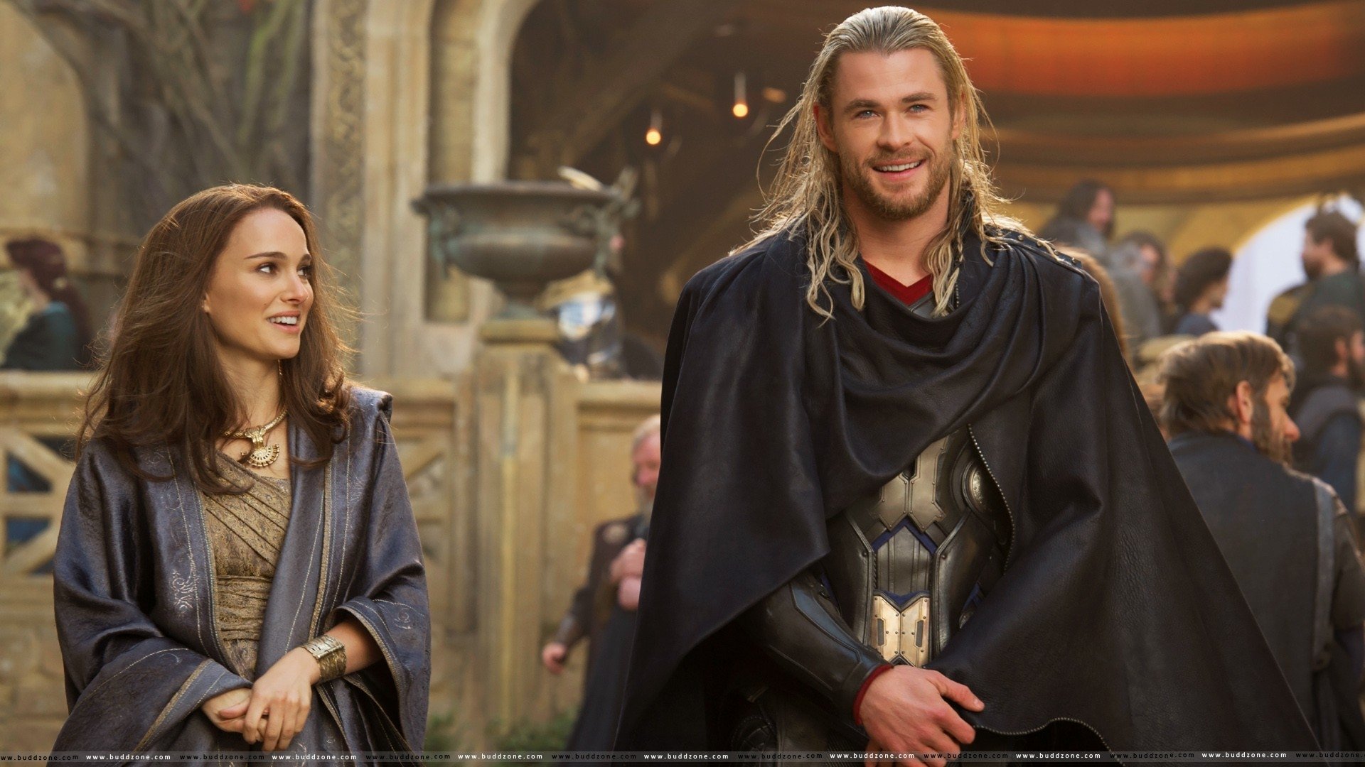 Actor playing Thor in armor and a dark cape smiles beside a woman in a grand hall — scene from the movie Thor: The Dark World.