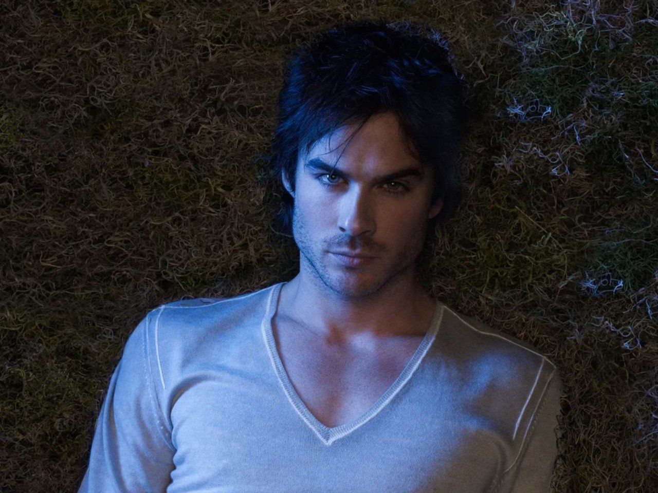 Ian Somerhalder gazes intensely while lying on grass, dressed in a light V-neck shirt, exuding a charismatic and mysterious aura.