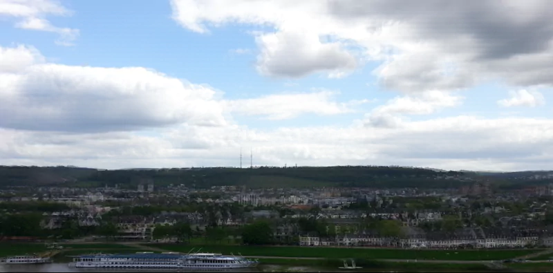 A panoramic view of a man-made city landscape under a cloudy sky, showcasing buildings, green spaces, and a riverboat along the water's edge.