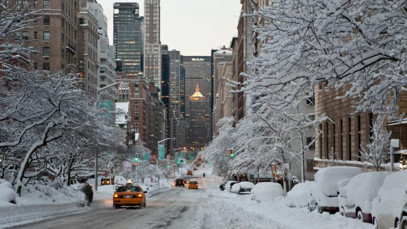 A snowy New York street lined with snow-covered trees and parked cars, with tall man-made buildings and a yellow taxi driving through the winter scene.