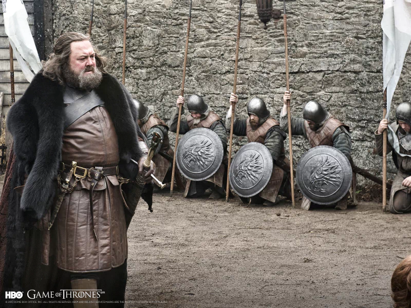 Scene from the TV show Game of Thrones: a bearded lord in furs stands in a courtyard while armed soldiers kneel with shields and spears against a stone wall.