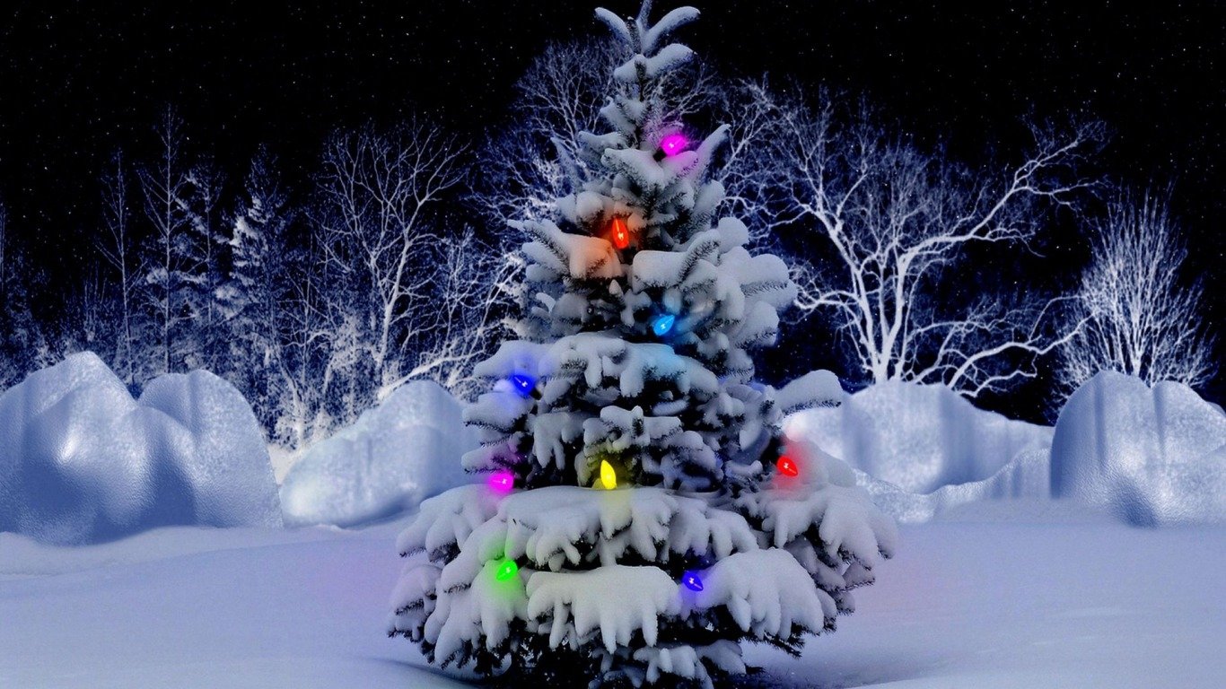 Snow-covered Christmas tree decorated with colorful lights stands glowing in a snowy holiday landscape at night.