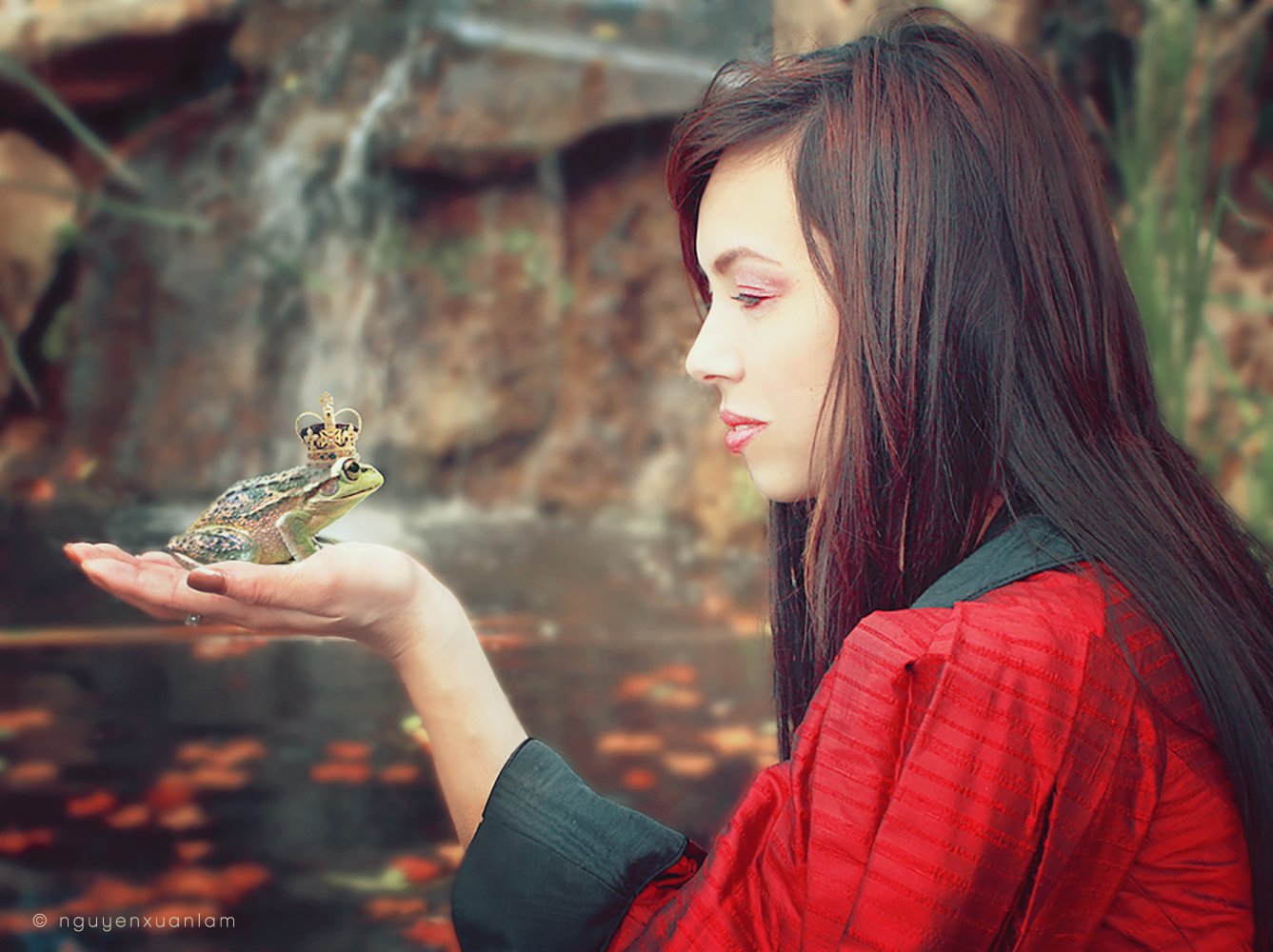 A fantasy-themed image of a woman in a red cloak holding a small frog in her hand, set against a natural, rocky background.