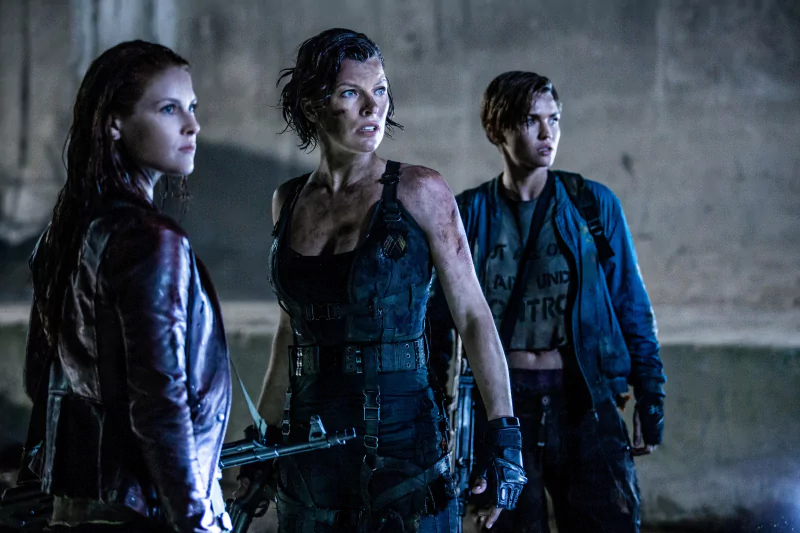 Battle-worn Alice (Resident Evil) in tactical gear stands flanked by two allies in a ruined, dim setting from the film Resident Evil: The Final Chapter.