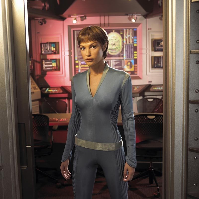 Crew officer in a form-fitting gray Starfleet uniform stands in a Star Trek: Enterprise ship interior, framed by a doorway with consoles and lit panels behind her.