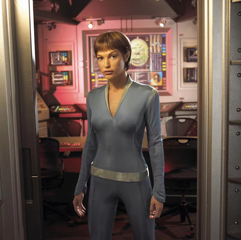 Crew officer in a form-fitting gray Starfleet uniform stands in a Star Trek: Enterprise ship interior, framed by a doorway with consoles and lit panels behind her.
